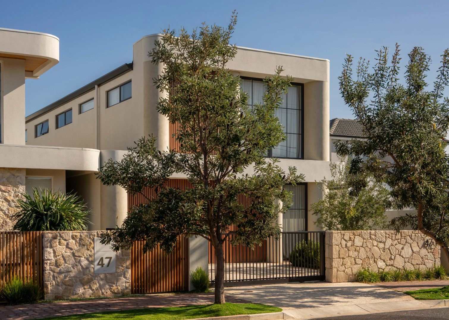 A contemporary two storey home behind a stone and timber front fence, with a street tree in front