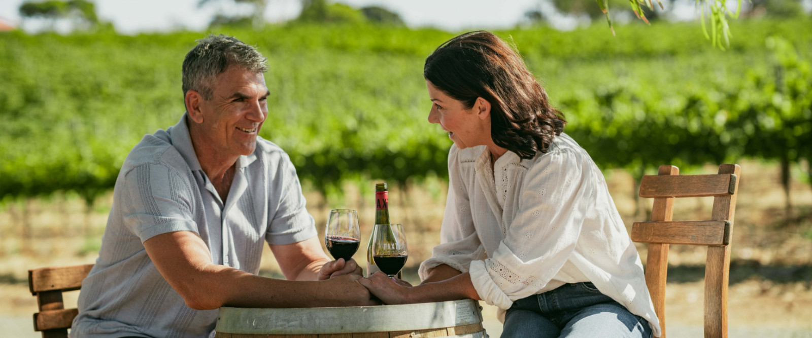 A couple seated at a wine barrel table between rows of vines